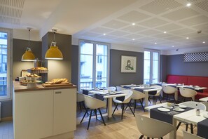 Un espace repas moderne, doté de tables et de chaises, d’un comptoir où sont présentés des aliments et de grandes fenêtres qui laissent entrer la lumière naturelle.