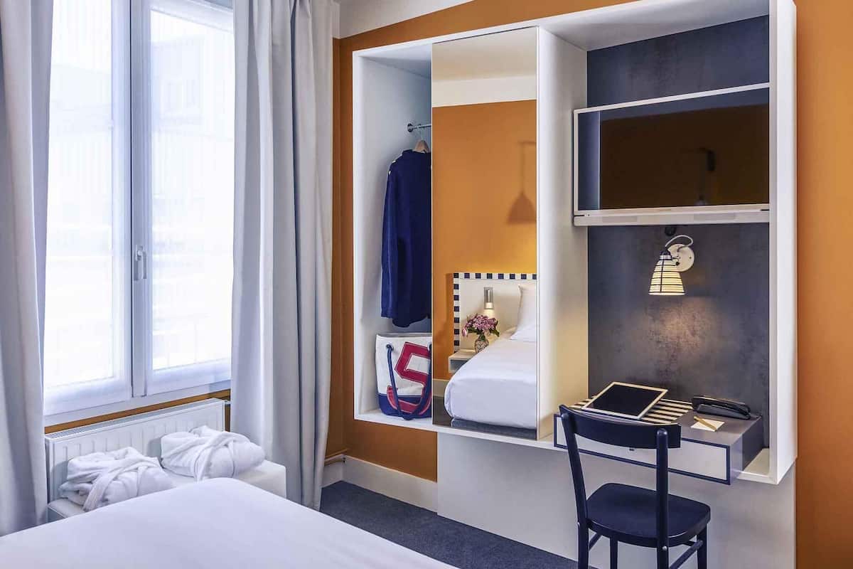 Une chambre d’hôtel avec un lit, un bureau et une chaise, un miroir et une fenêtre avec des rideaux.