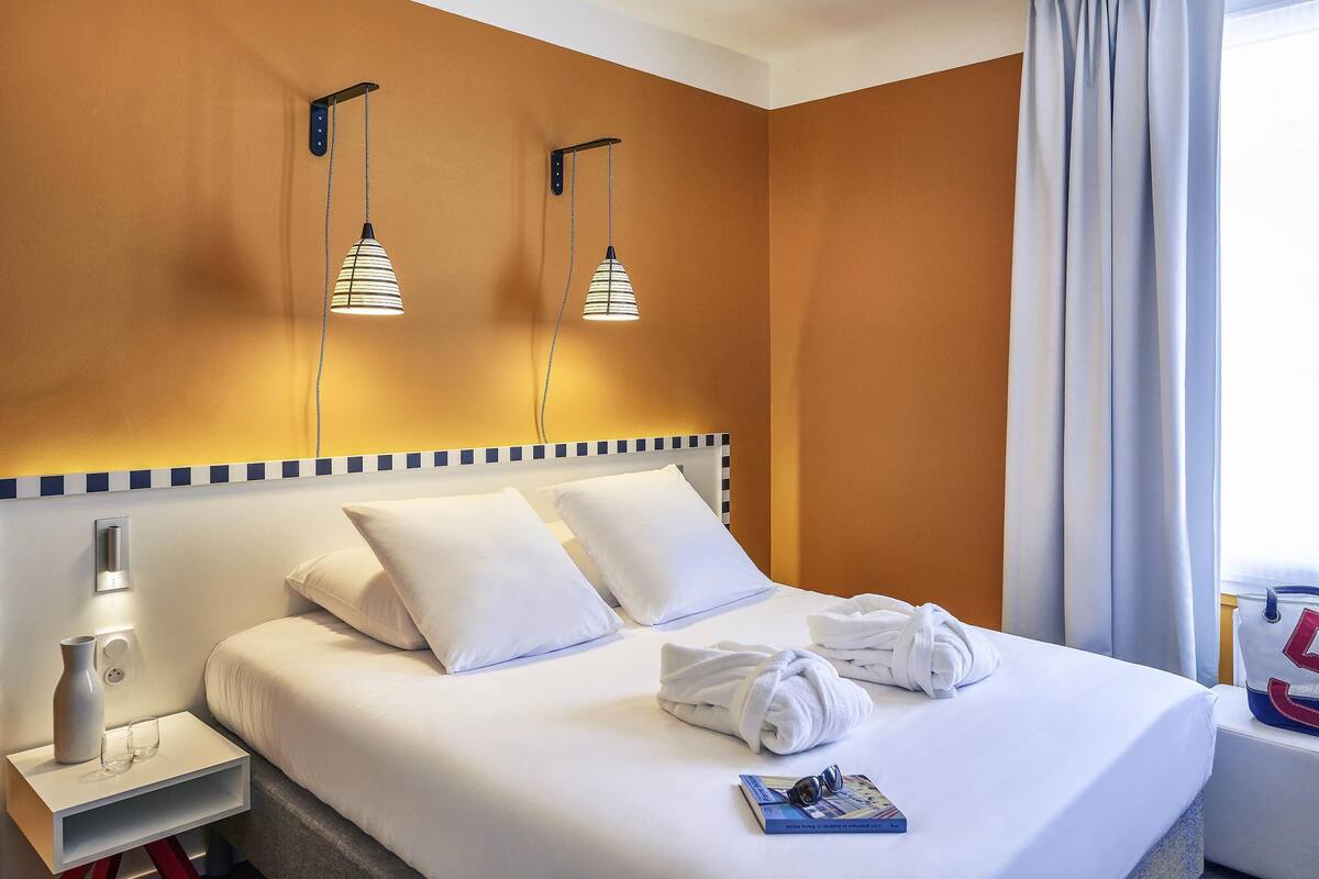 Una habitación de hotel con una cama bien hecha, dos lámparas de noche, una mesita de noche con un florero y vasos, y una ventana con cortinas.
