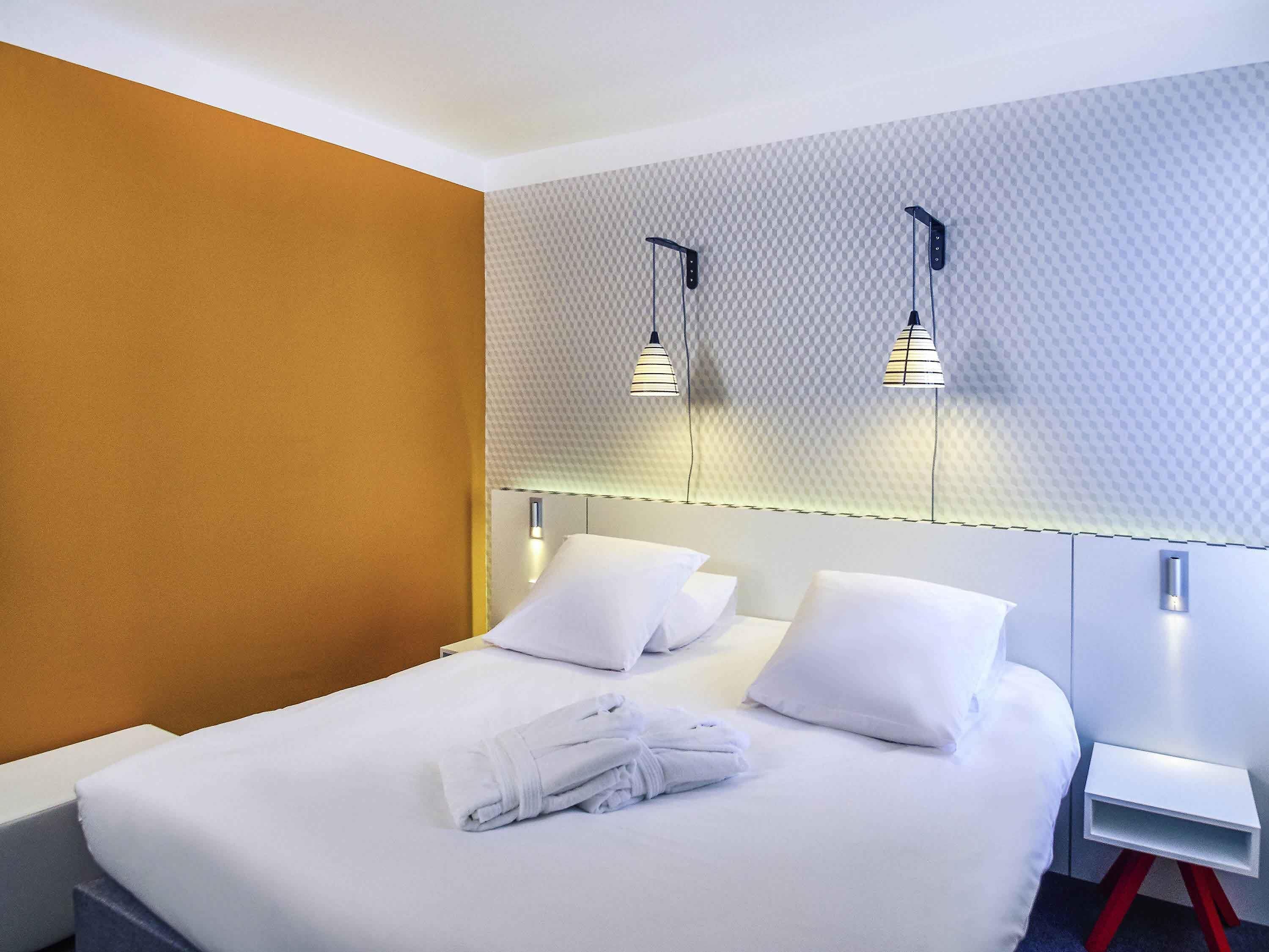 Ein Hotelzimmer mit einem ordentlich bezogenen Bett, weißen Kissen und einem gefalteten Handtuch. Es gibt zwei Nachttische, einer mit rotem und einer mit weißem Fuß. Der Raum hat eine strukturierte Wand und eine Decke mit einer Leuchte.
