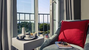 Une chambre moderne avec une grande fenêtre, une table de chevet blanche sur laquelle se trouve un plateau avec des articles pour le petit-déjeuner, un fauteuil gris avec un coussin rouge et un livre posé sur l’accoudoir.