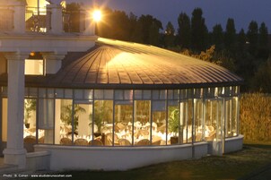 Estructura con paredes de vidrio que contiene mesas y sillas, probablemente un comedor o sala de estar, con exterior blanco y techo metálico.