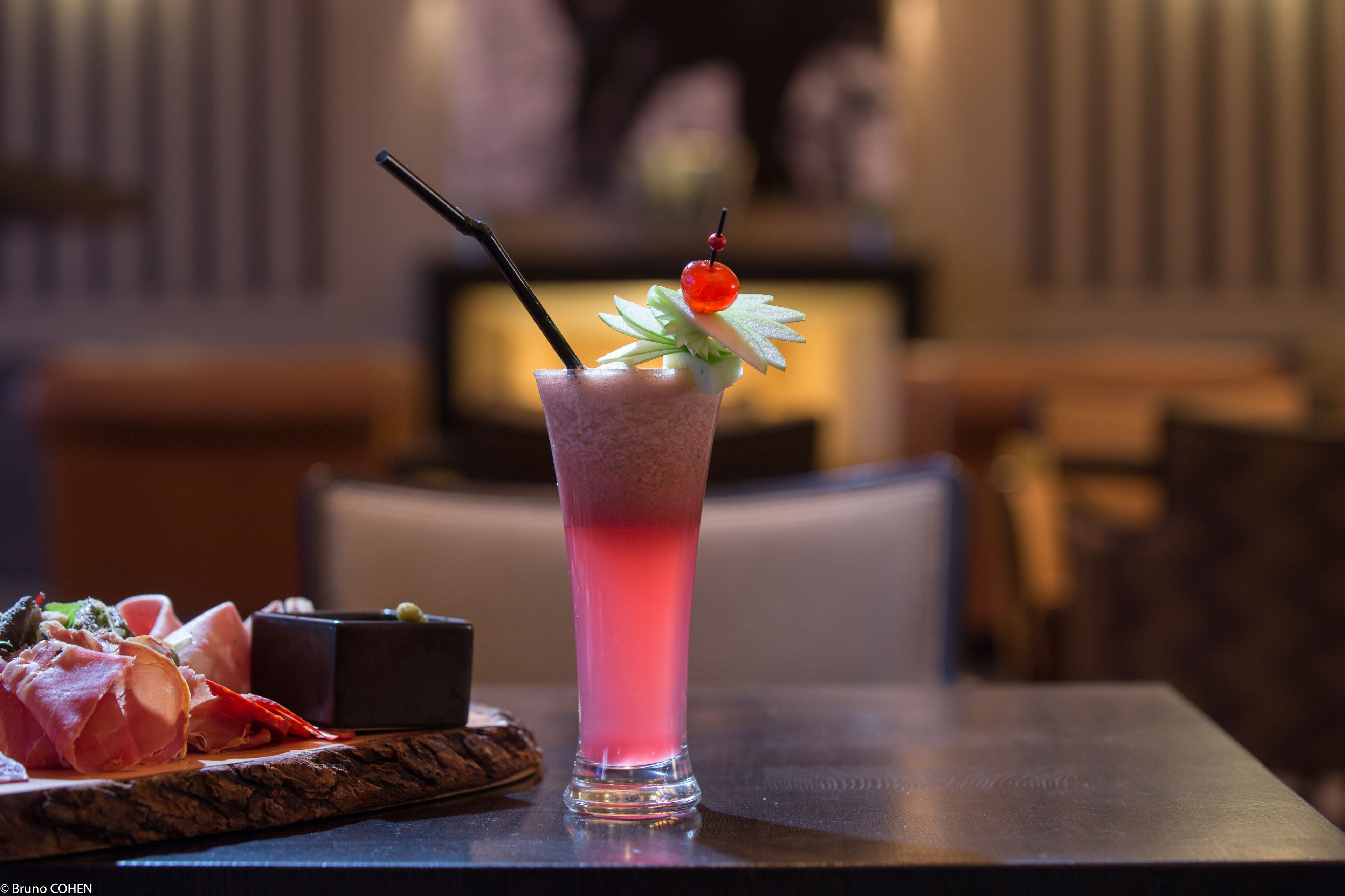 A cocktail with a cherry garnish and a straw, placed on a table with a blurred background.