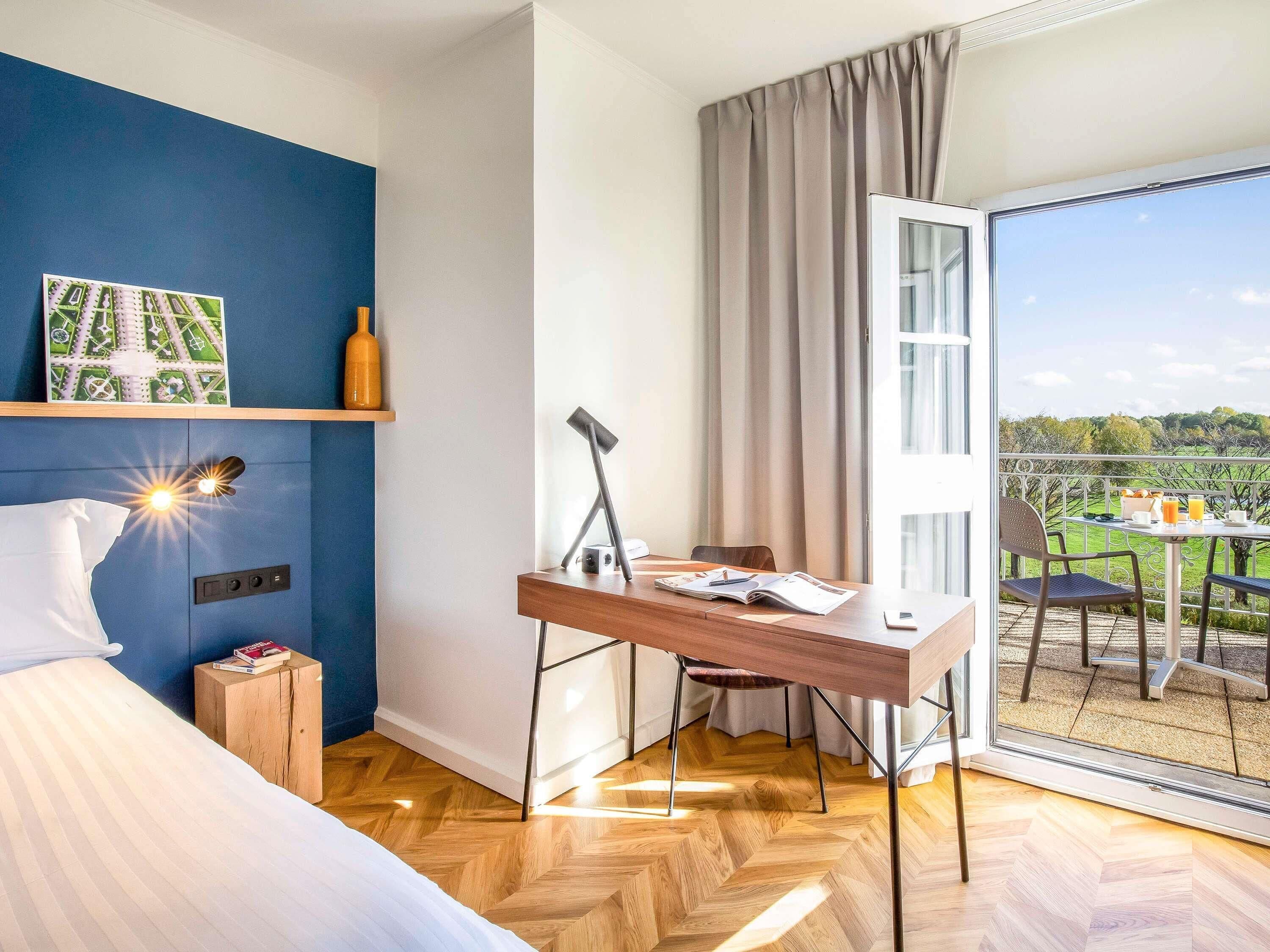 A hotel room with a bed, desk, and a view of a balcony with outdoor seating.