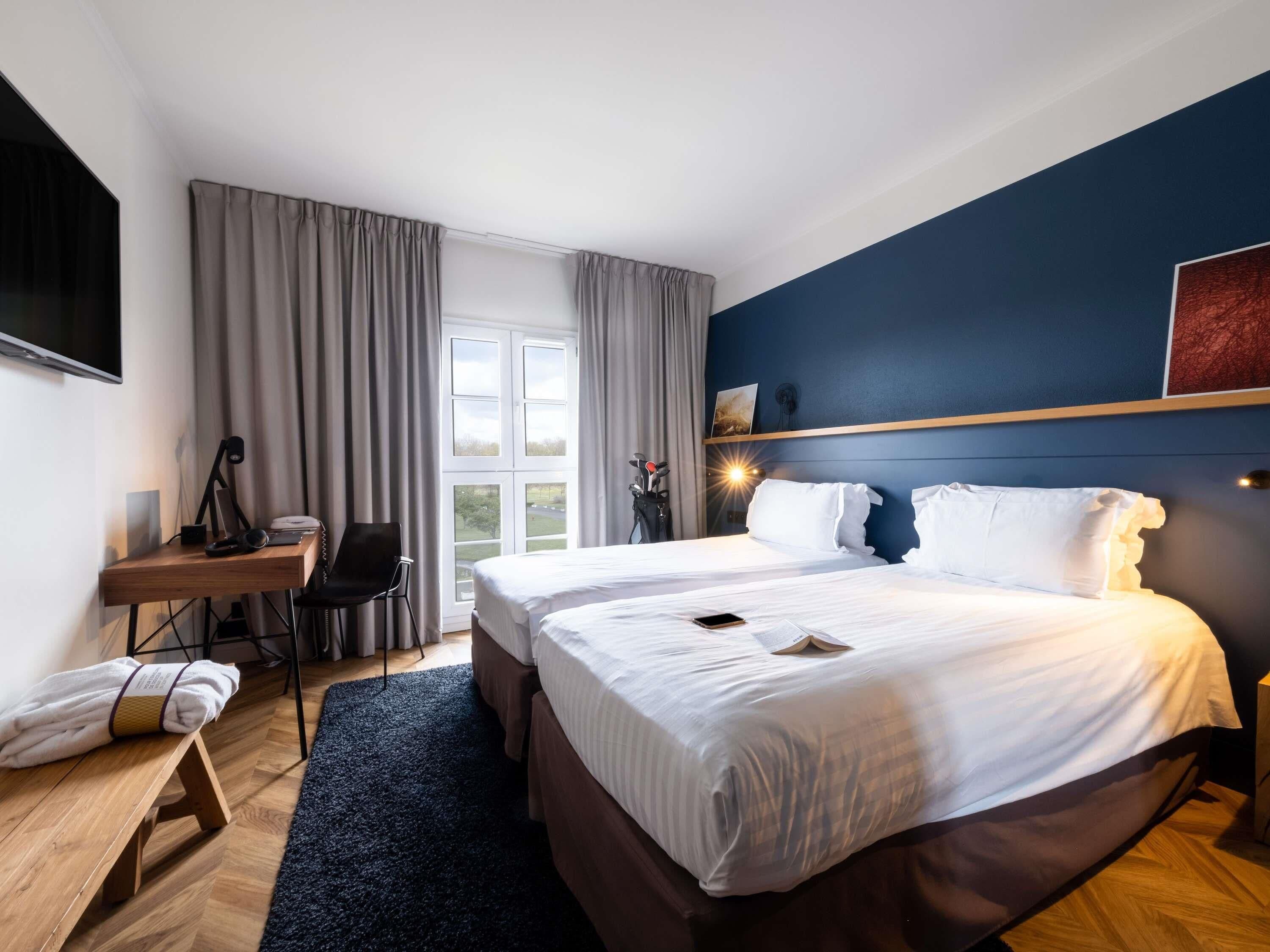 A hotel room with a bed, a desk, a chair, a TV, and a window with curtains.
