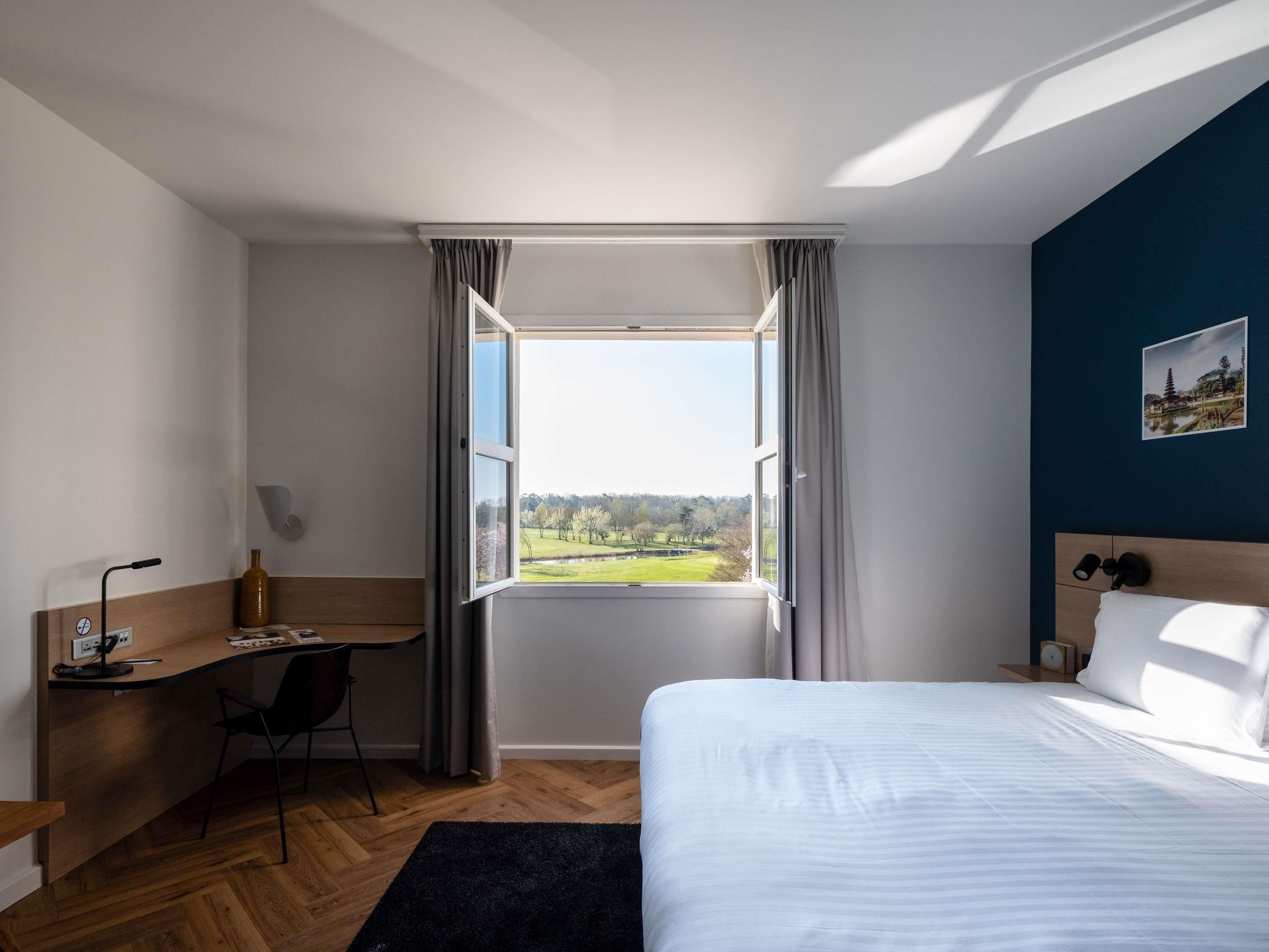 A hotel room with a bed, a desk, a chair, a window with curtains, and a view of a green landscape.