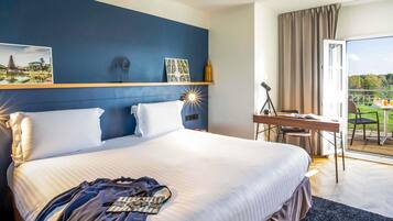 Habitación de hotel con una cama grande, un escritorio con una silla, un balcón con mesa y sillas, y vistas al exterior.