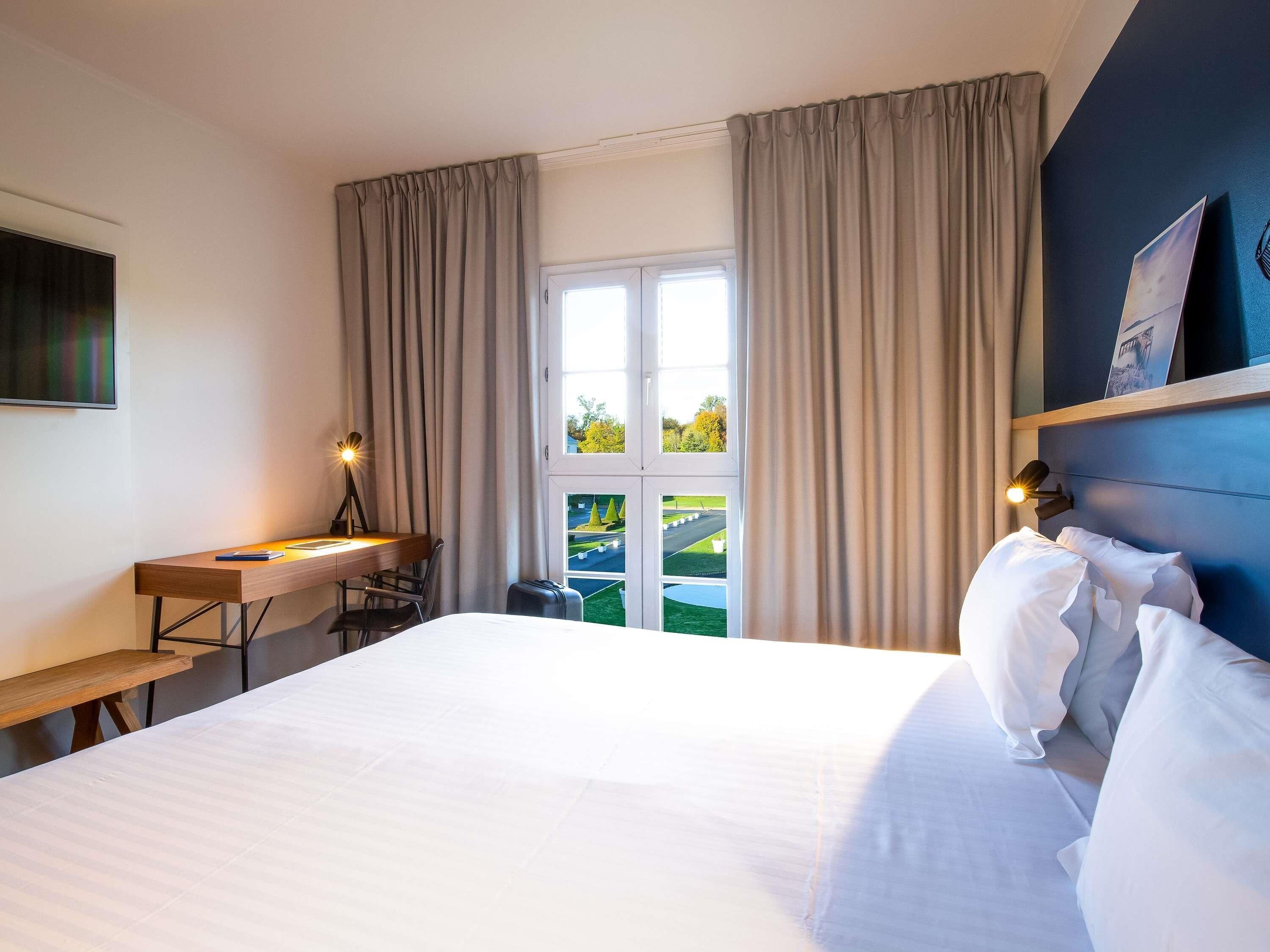A hotel room with a bed, desk, chair, and a window with curtains.