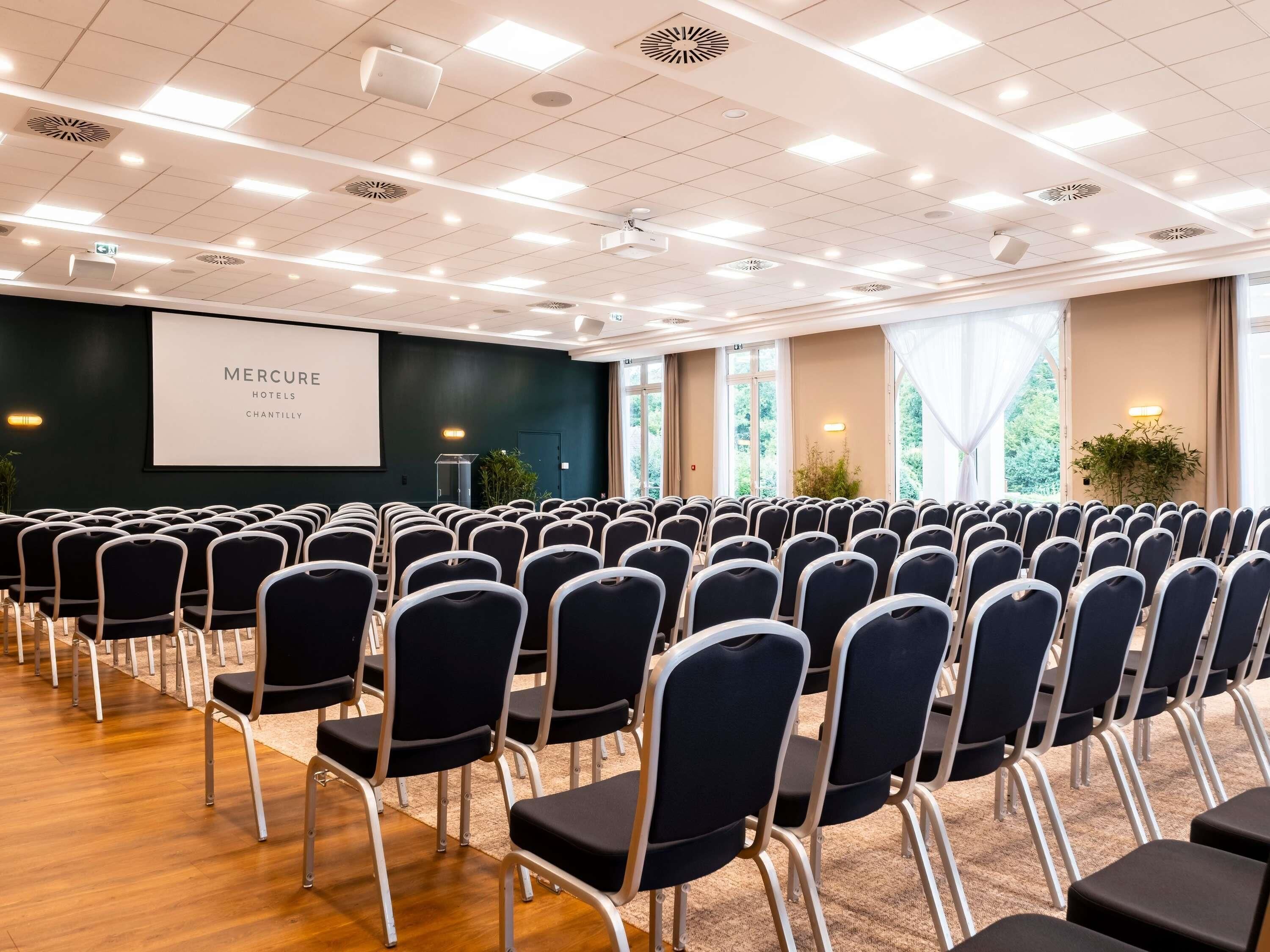 Une salle de conférence avec des rangées de chaises, un écran de projection et un pupitre.