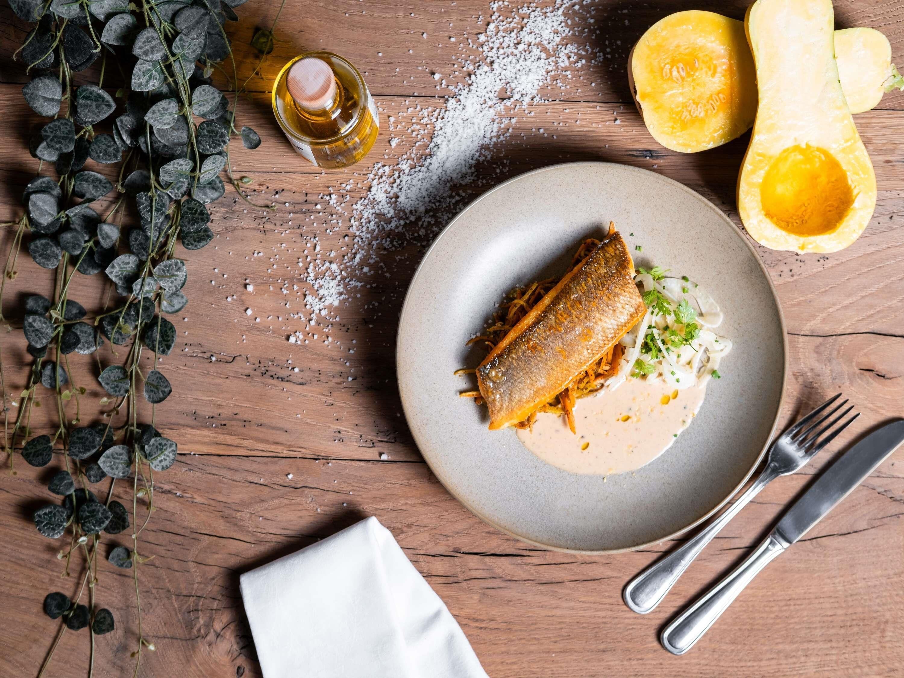 A plate with a fish fillet, a bowl of sauce, and a butternut squash half on a wooden table.