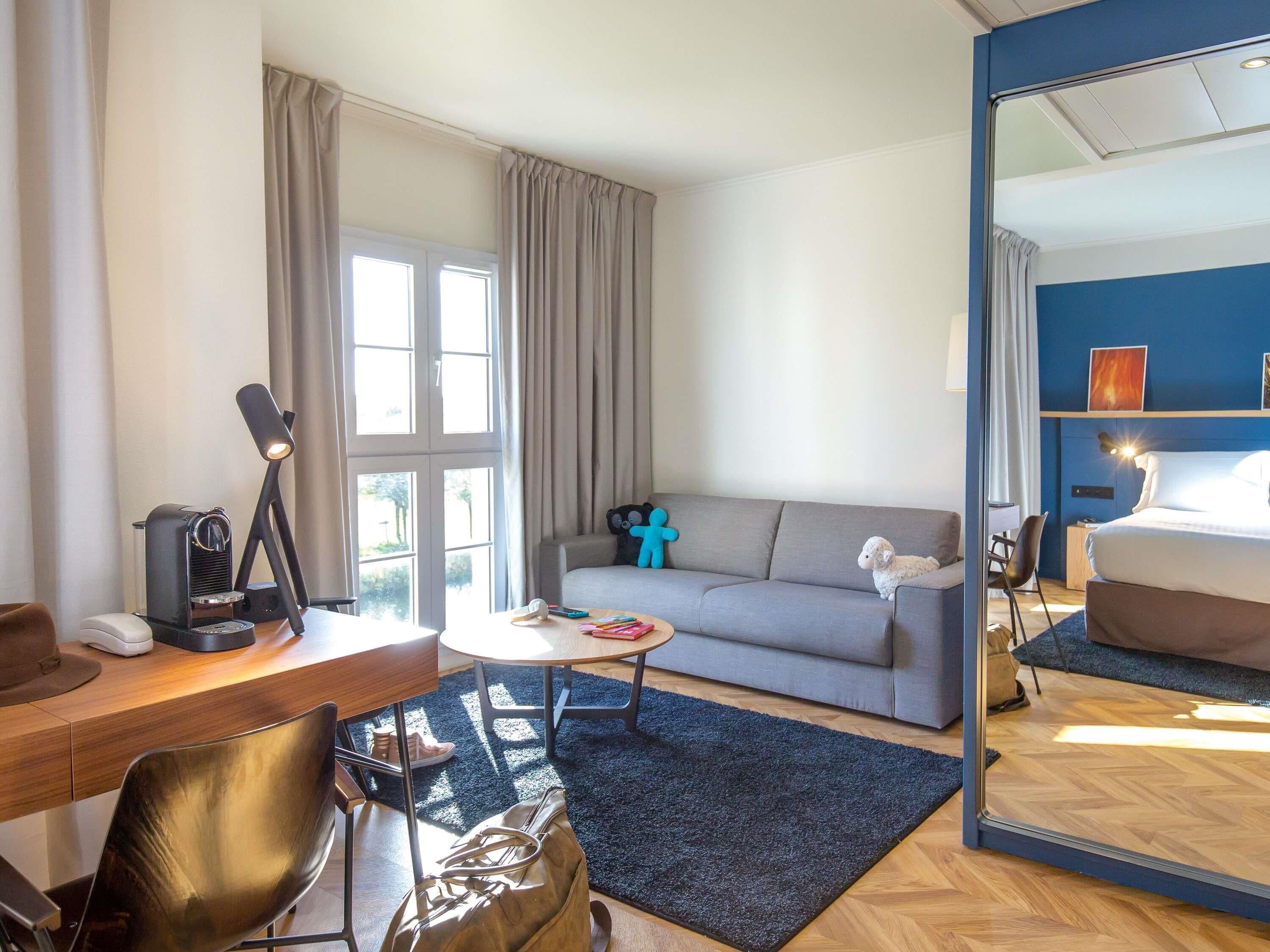 A hotel room with a desk, sofa, and a large mirror reflecting the bed.
