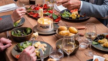 Un gruppo di persone che cena a un tavolo di legno, con diversi piatti, tra cui una casseruola di formaggio fuso, insalate e pane.