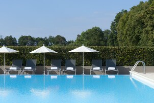 Piscina con sillas de descanso y sombrillas, rodeada de setos y árboles.