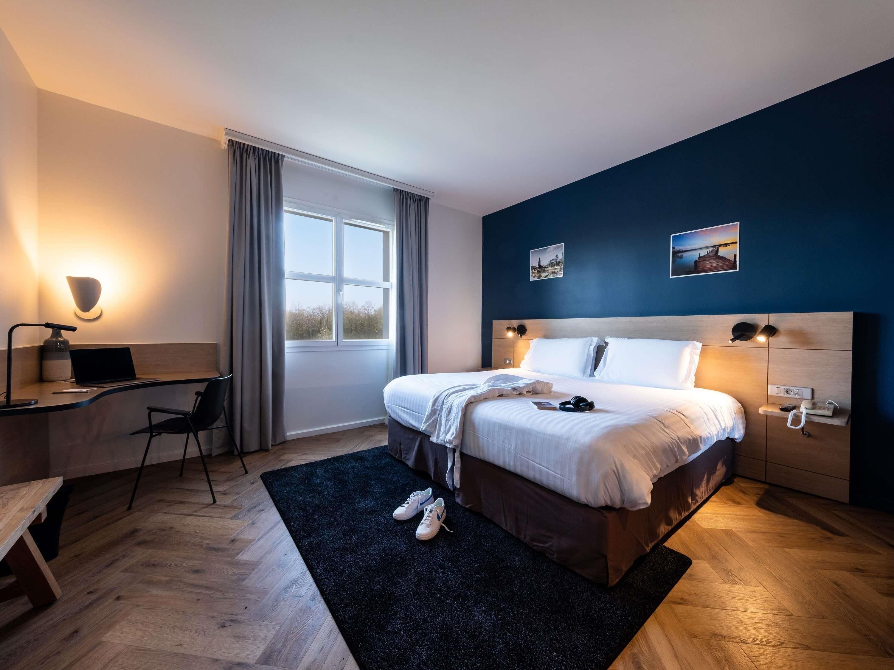 Habitación de hotel moderna con una cama grande, un escritorio, una silla y un ventanal con cortinas.