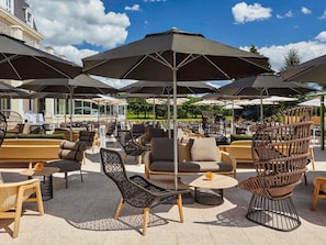 Outdoor seating with umbrellas, wicker furniture, and a view of a building in the background. - Mercure Chantilly Resort & Conventions (Vineuil-Saint-Firmin)
