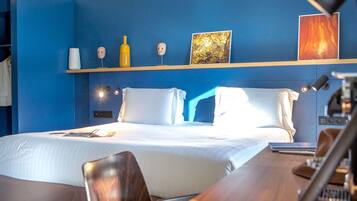 Habitación de hotel con cama, mesitas de noche, escritorio y una silla. Se observan estantes en la pared con objetos decorativos.