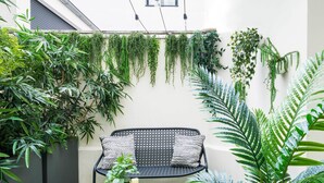 Un piccolo patio recintato con una panca in metallo nero, un tavolino con candele e diverse piante verdi.