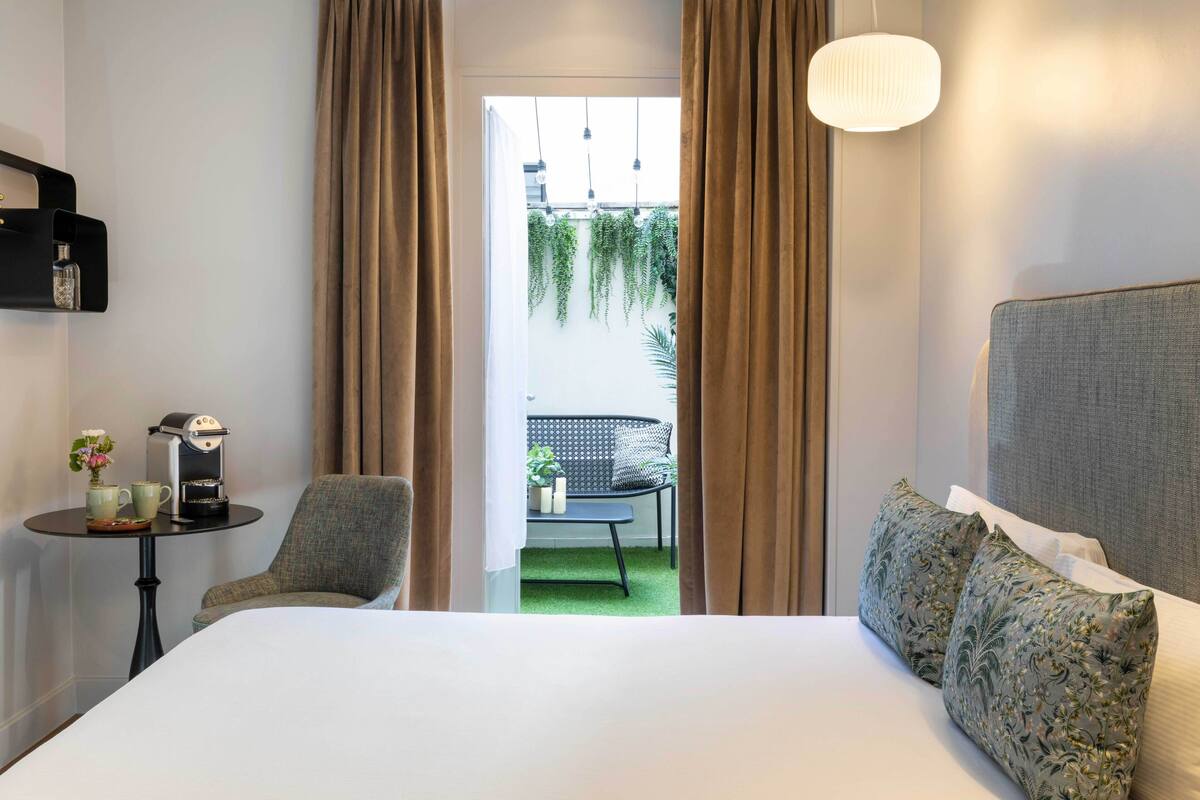 A hotel room with a large bed, a small table with a coffee maker and cups, a chair, and a view of a balcony with plants and a chair.