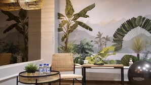 Una stanza con una sedia in vimini, un tavolino con bottiglie d'acqua, una pianta decorativa e un grande murale con paesaggi tropicali.