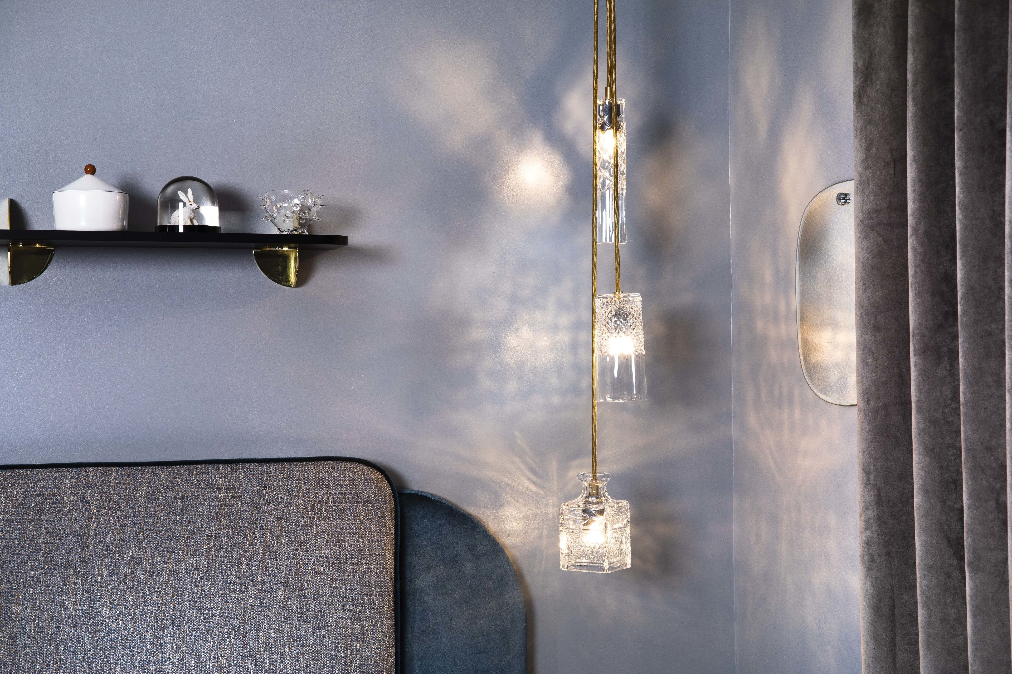 A modern interior with a wall-mounted shelf holding decorative items, a hanging crystal light fixture, and a partial view of a grey upholstered chair.