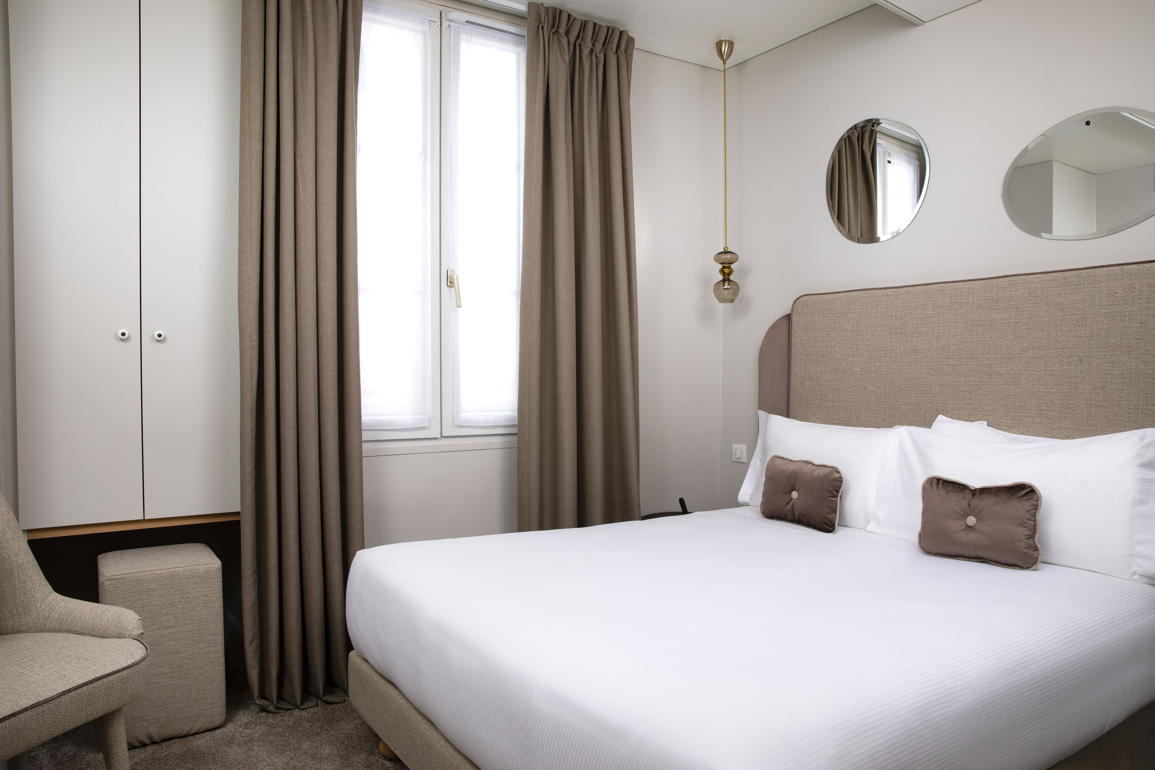 A hotel room with a bed, a chair, a wardrobe, and a window with curtains.