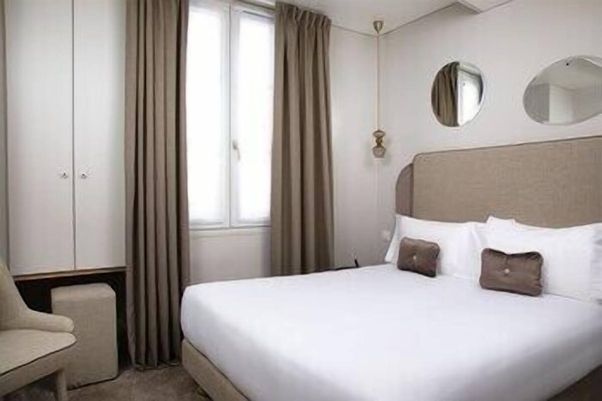 A hotel room with a bed, a chair, a wardrobe, and a window with curtains.
