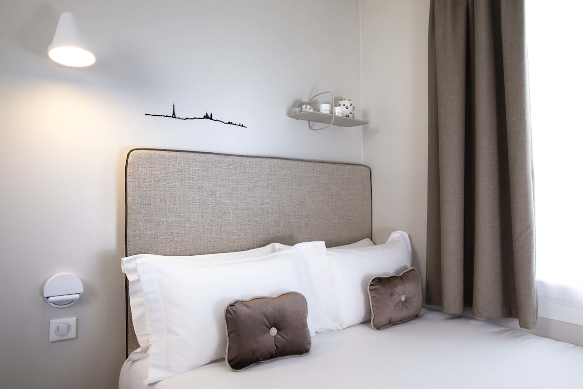 A neatly made bed with white and brown pillows, a wall-mounted shelf with toiletries, a wall clock, and a window with curtains.