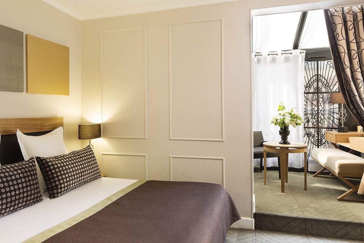 A hotel room with a bed, a nightstand with a lamp, a small table with a vase of flowers, and a view of the outside balcony.