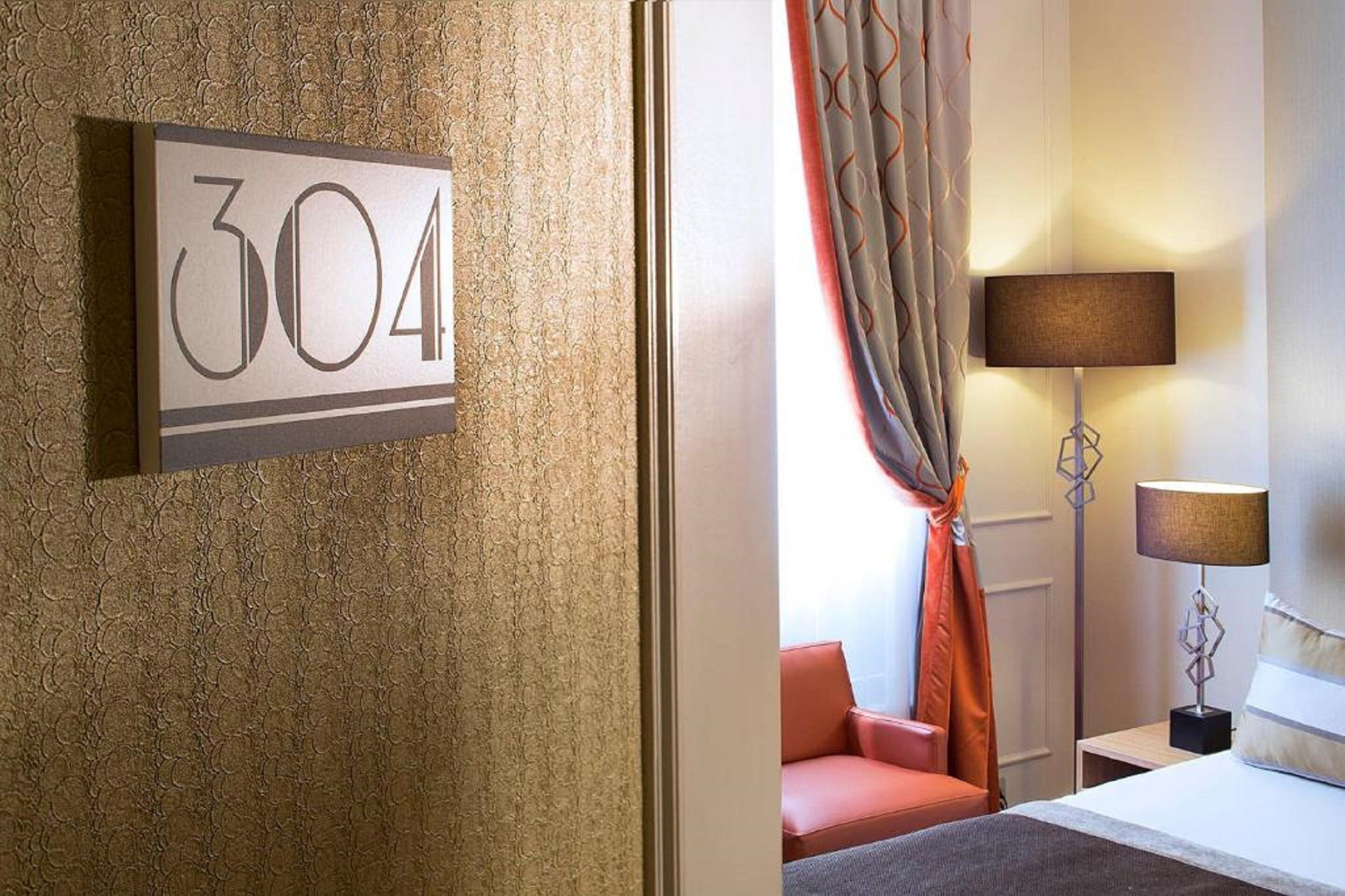A hotel room with a number sign, a bed, a nightstand, a lamp, and a window with curtains.