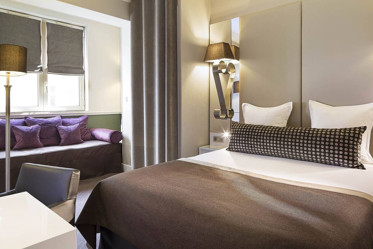 A hotel room with a large bed, a bedside table, a lamp, a window with blinds, and a seating area with cushions.