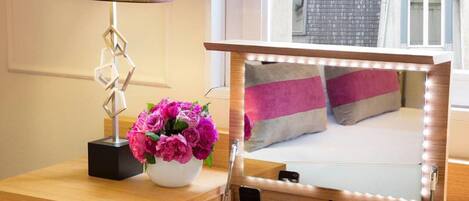 A hotel room with a vanity area, a mirror with lights, a vase with flowers, and a bed with pillows.