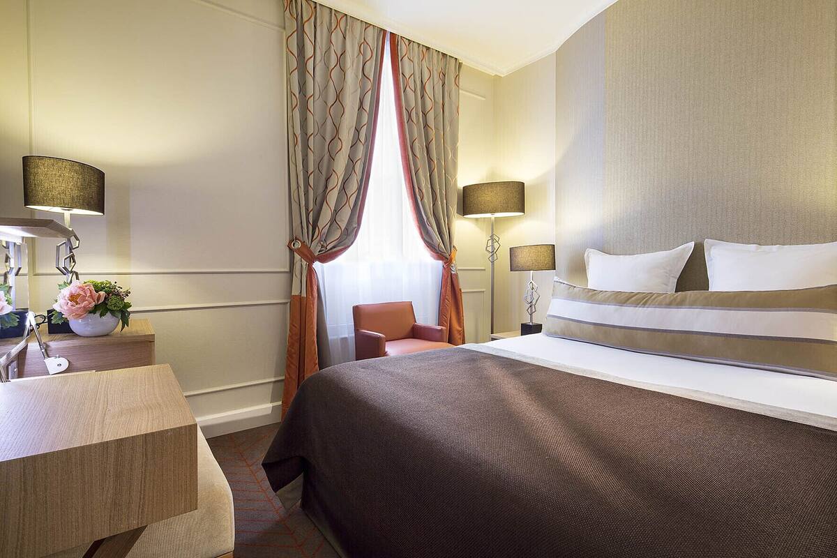 A hotel room with a large bed, a desk with a lamp, a chair, and a window with curtains.