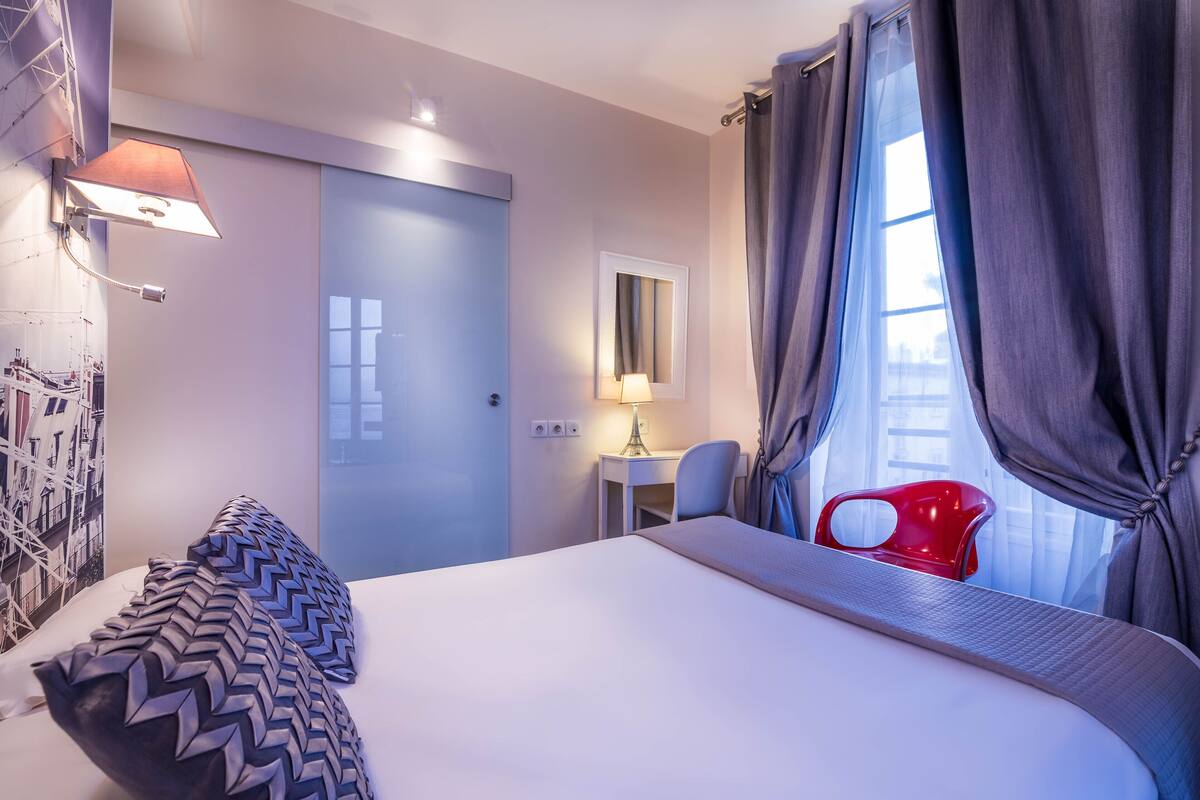 Una habitación de hotel con cama, escritorio, silla, lámpara y ventana con cortinas.