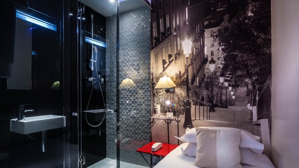 Una habitación de hotel moderna con cabina de ducha de vidrio, una mesita de noche con lámpara y una cama con almohadas.