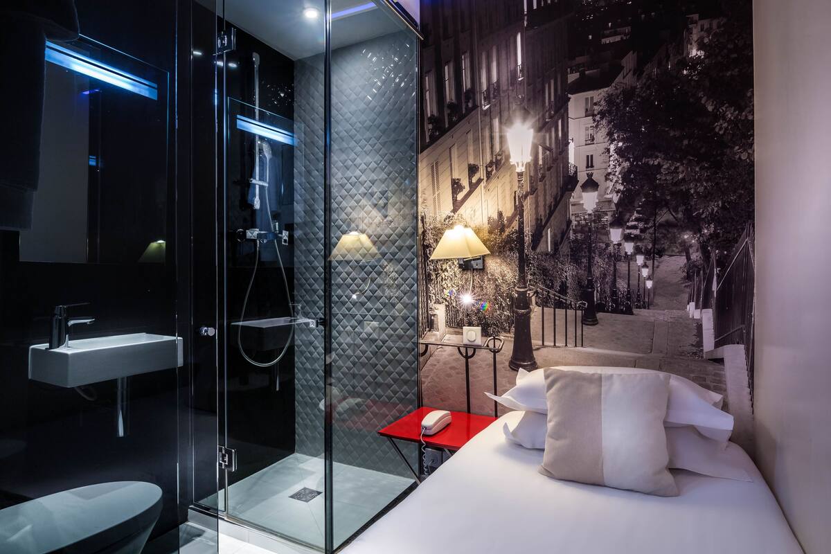 Una habitación de hotel moderna con cabina de ducha de vidrio, una mesita de noche con lámpara y una cama con almohadas.