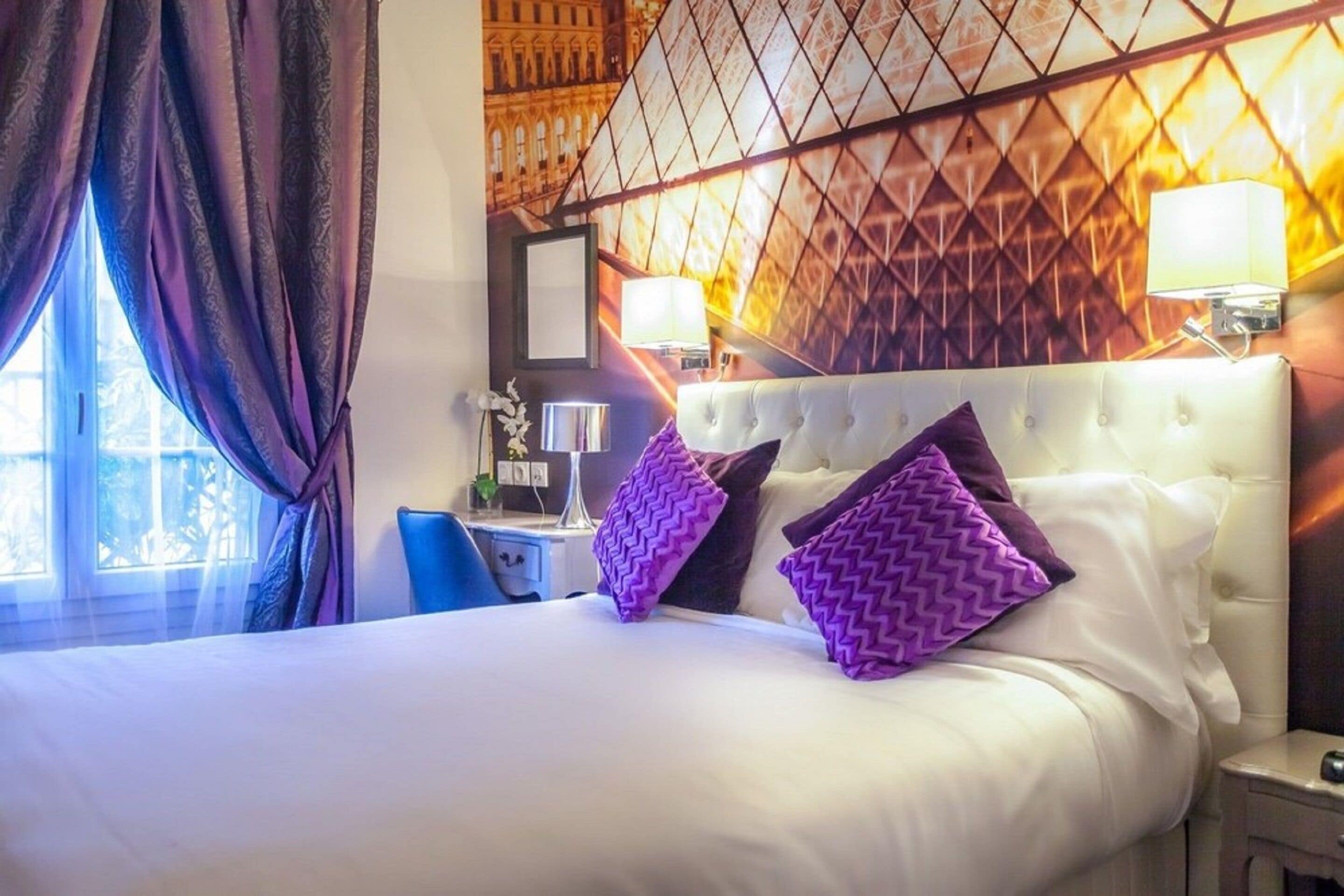 Habitación de hotel con una cama grande, dos almohadas moradas, una mesita de noche con lámpara y una ventana con cortinas.
