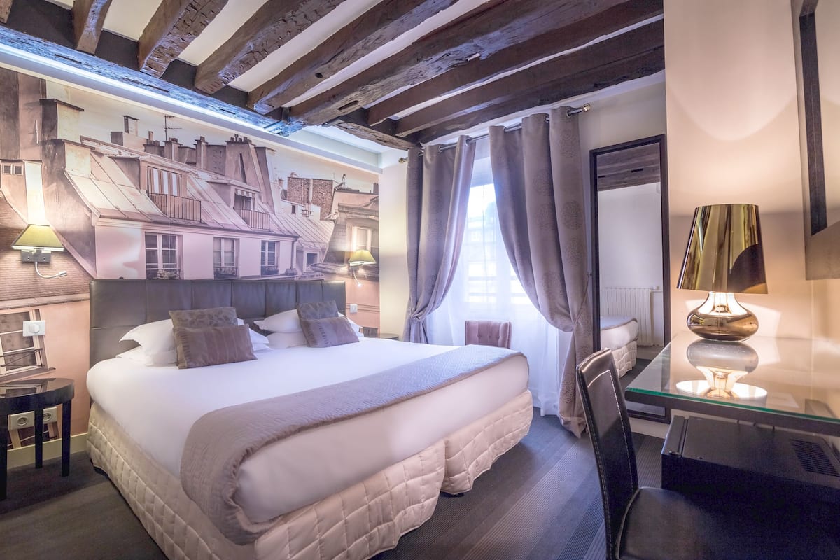 Habitación de hotel con una cama grande, un escritorio con lámpara y un mural con edificios en la pared.