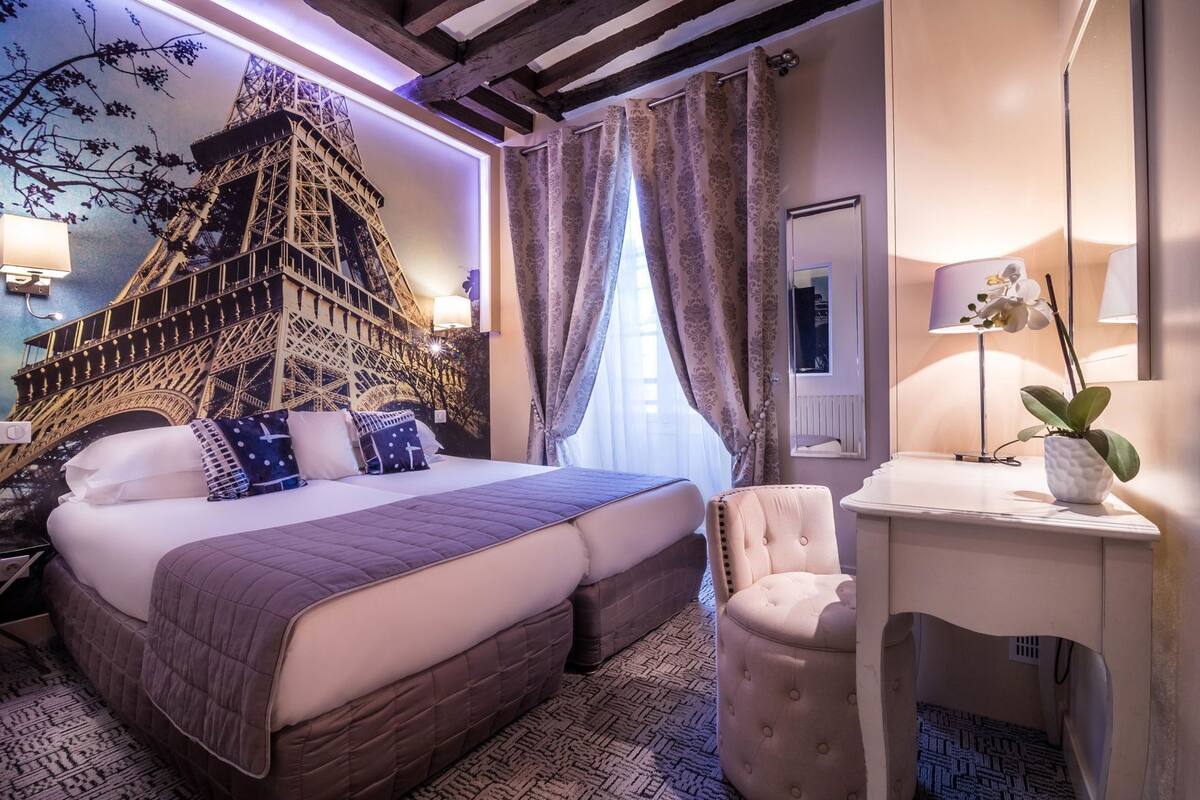 Habitación de hotel con cama, una silla, un escritorio, un espejo y una pared con un mural decorativo de la Torre Eiffel.