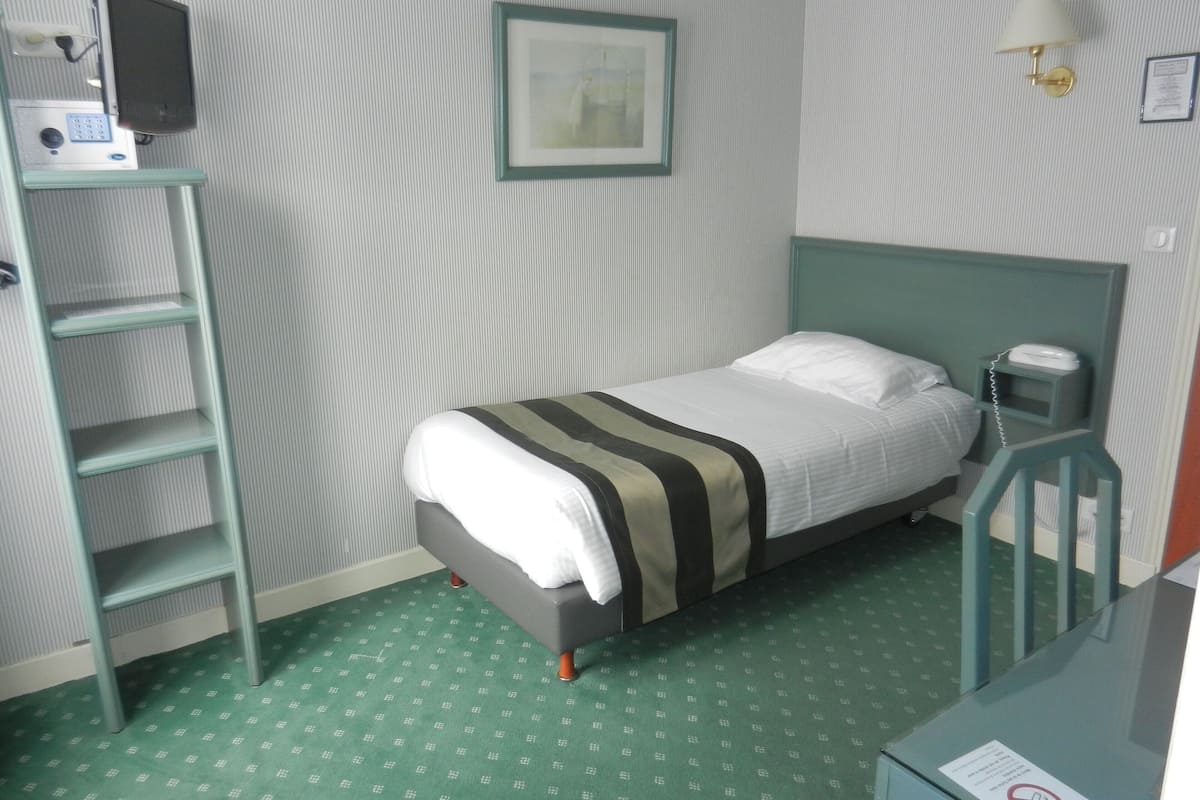 A hotel room with a bed, a desk, a chair, a telephone, a lamp, a picture on the wall, and a TV on a stand.