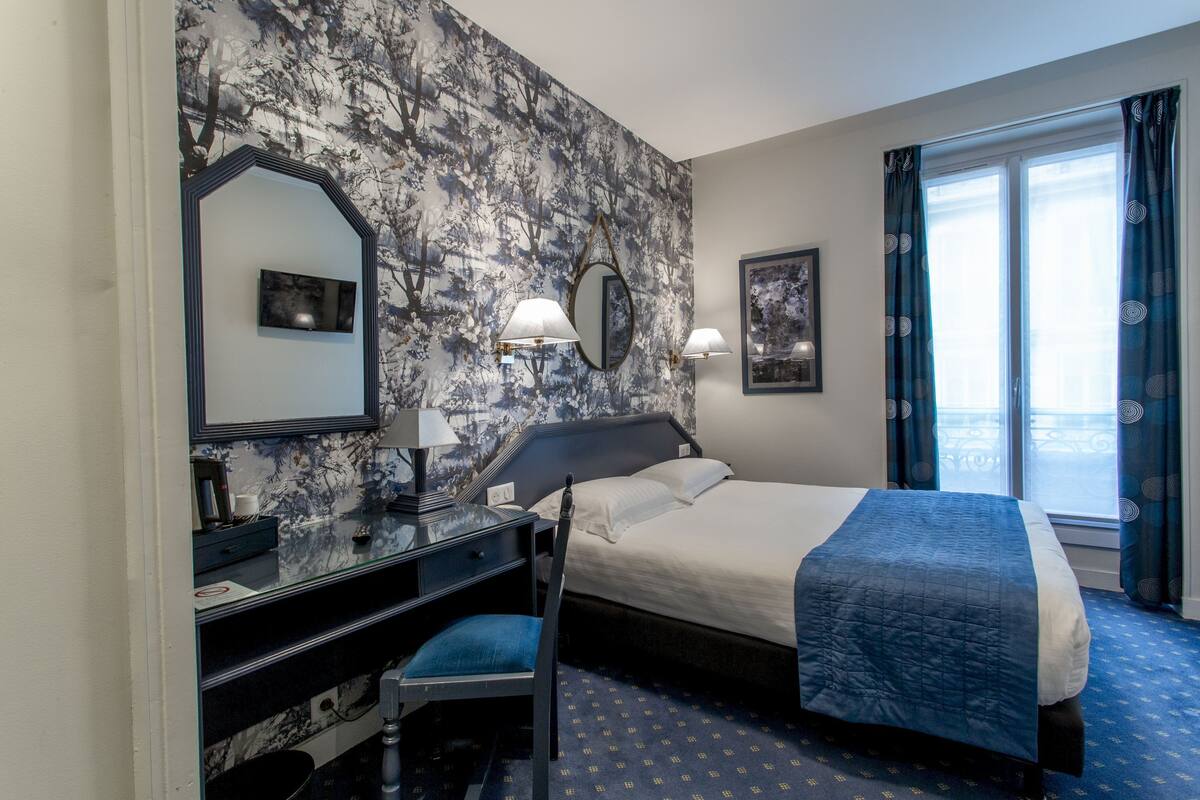 A hotel room with a bed, a desk with a chair, a mirror, and a window with curtains.