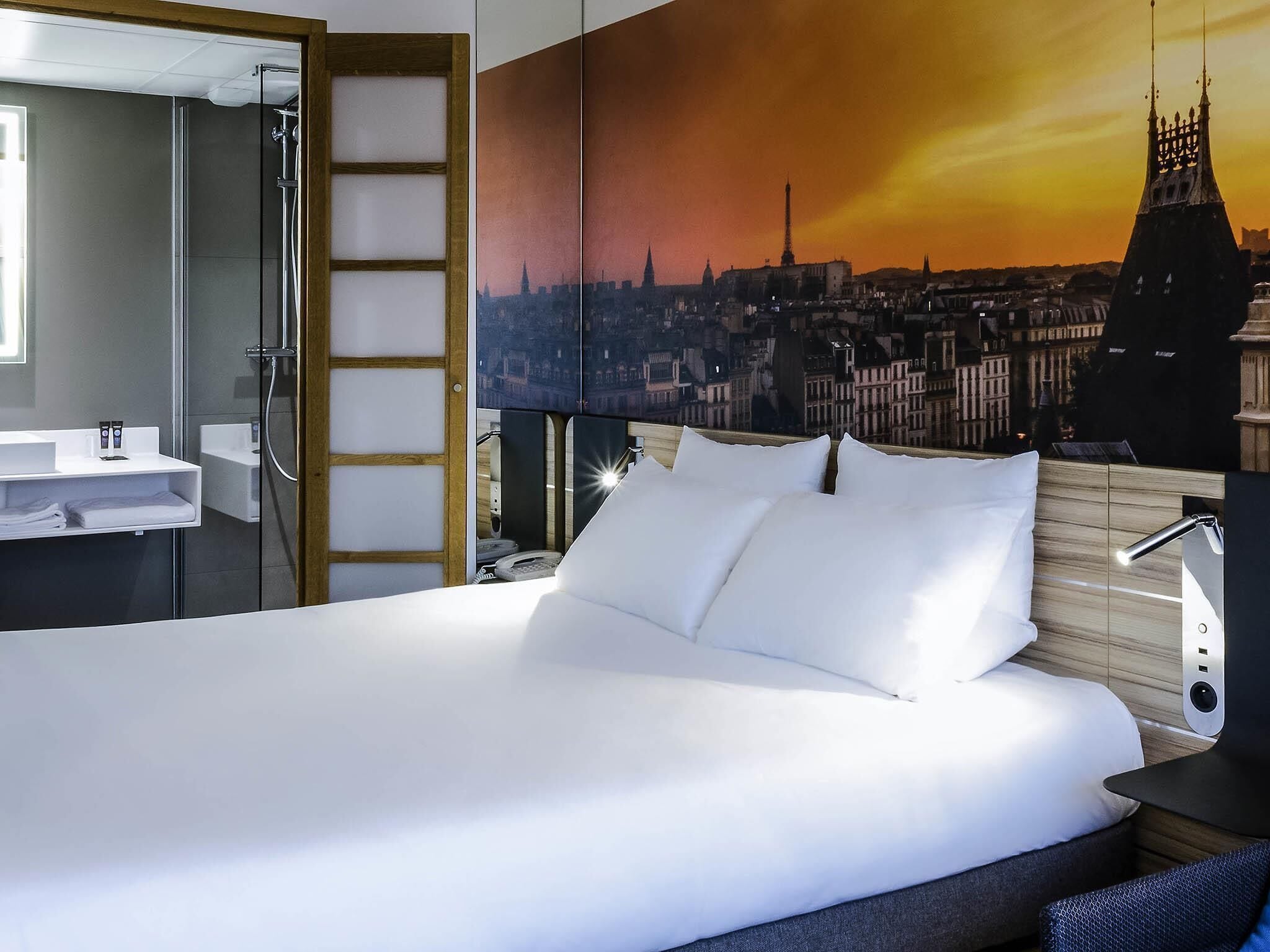 A hotel room with a bed, a bathroom with a shower, and a cityscape mural on the wall.