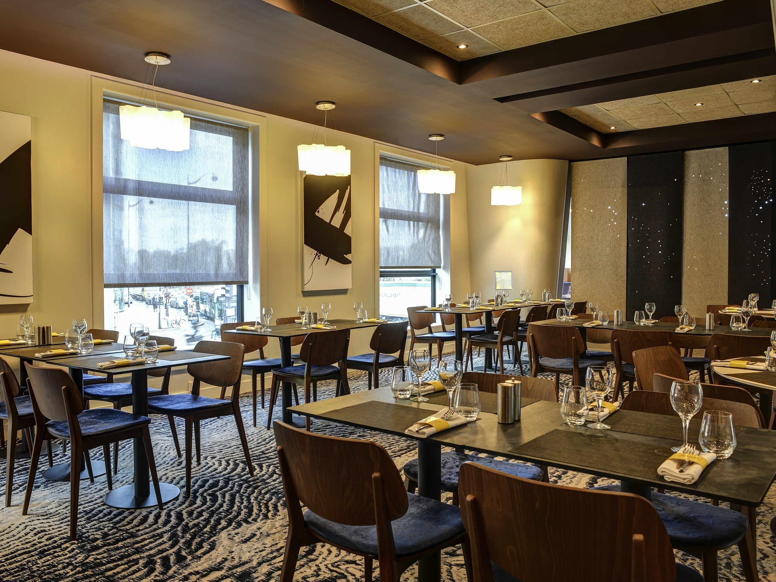 A hotel dining area with multiple tables, chairs, and large windows.