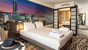 Ein Hotelzimmer mit Blick auf die Stadt, ein Bett mit weißen Leinentüchern, eine Sitzecke mit Hut und Sonnenbrille sowie ein gemustertes Teppich.