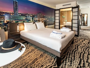 Une chambre dâhĂŽtel avec vue sur la ville, un lit avec du linge de maison blanc, un coin salon avec un chapeau et des lunettes de soleil, et une moquette Ă motifs.