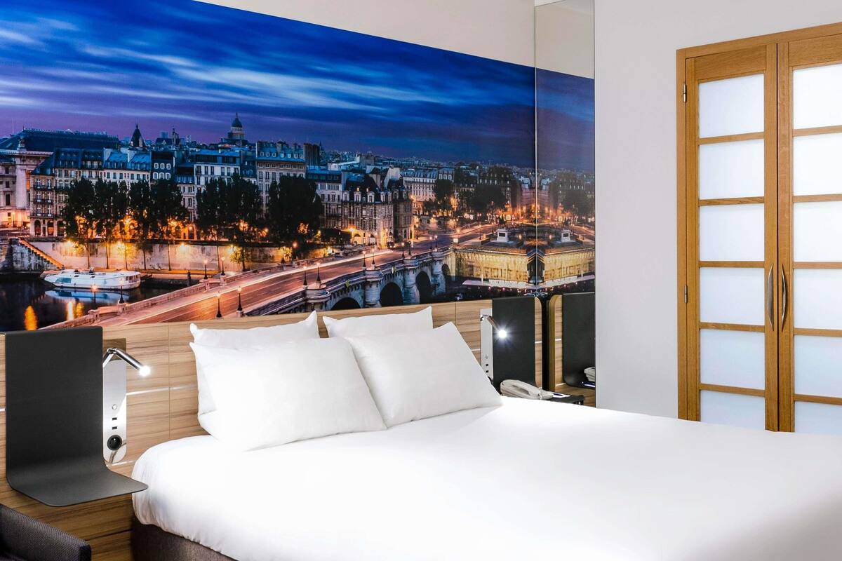 Une chambre d’hôtel avec un grand lit, une table de chevet, une lampe et une vue sur le paysage urbain illuminé la nuit.
