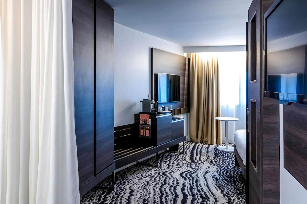 Une chambre d’hôtel moderne dotée d’une grande armoire, d’un téléviseur à écran plat, d’un minibar et d’un tapis à motifs.