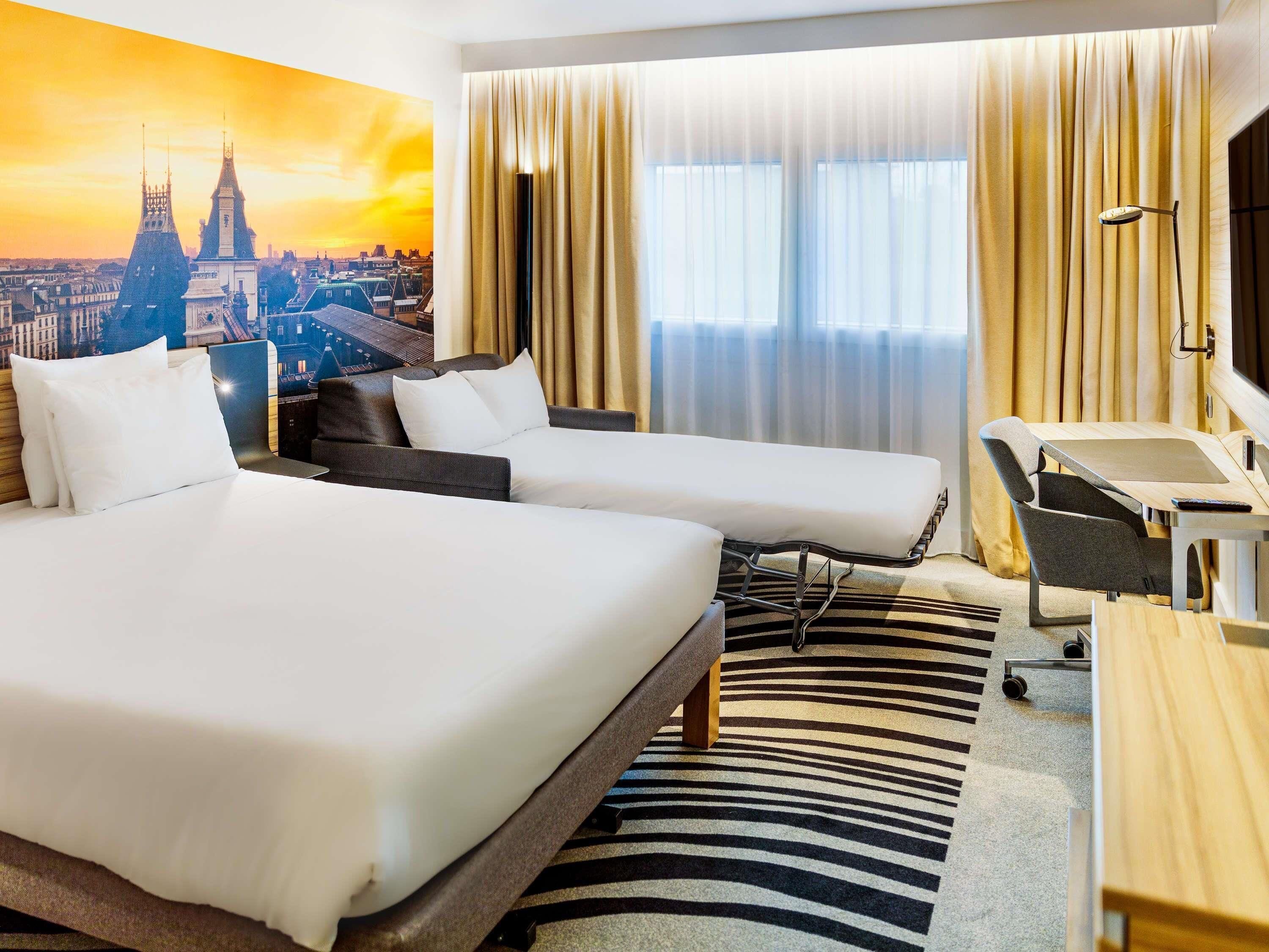 A hotel room with two beds, a desk, a chair, a zebra-patterned rug, and a cityscape view.