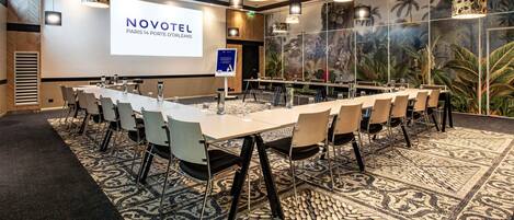 A conference room with a long table, chairs, a projector screen, and a tropical mural.