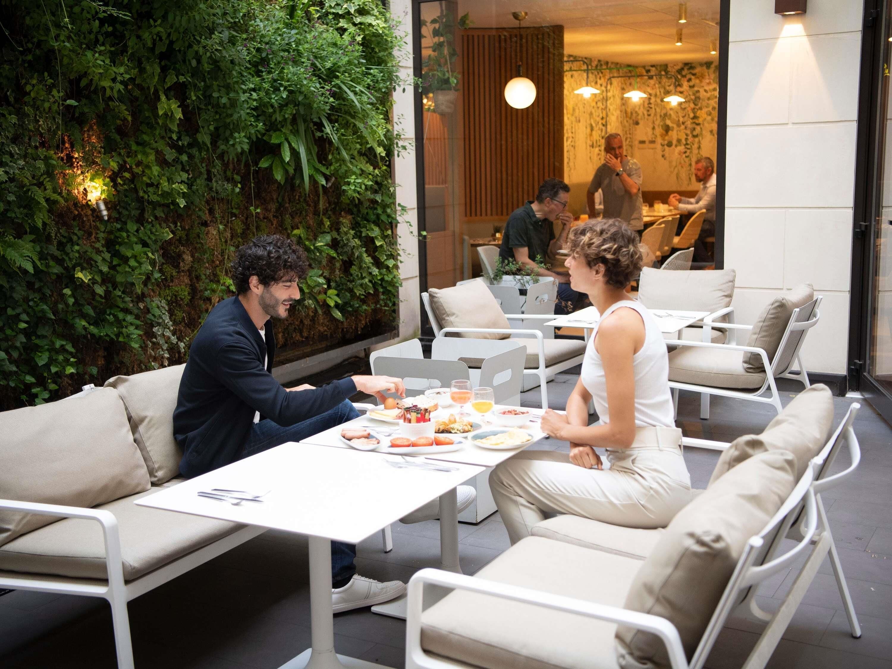 Un espacio de comedor al aire libre con mesas y sillas, rodeado de follaje y un interior moderno.
