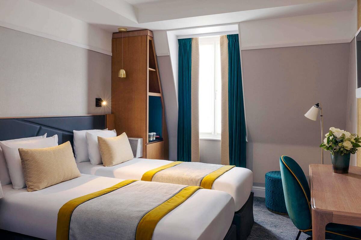 A hotel room with two beds, a desk, a chair, a lamp, and a window with curtains.